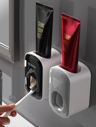Two wall-mounted ECOCO automatic toothpaste dispensers in black and gray, shown dispensing toothpaste onto a toothbrush in a modern bathroom.